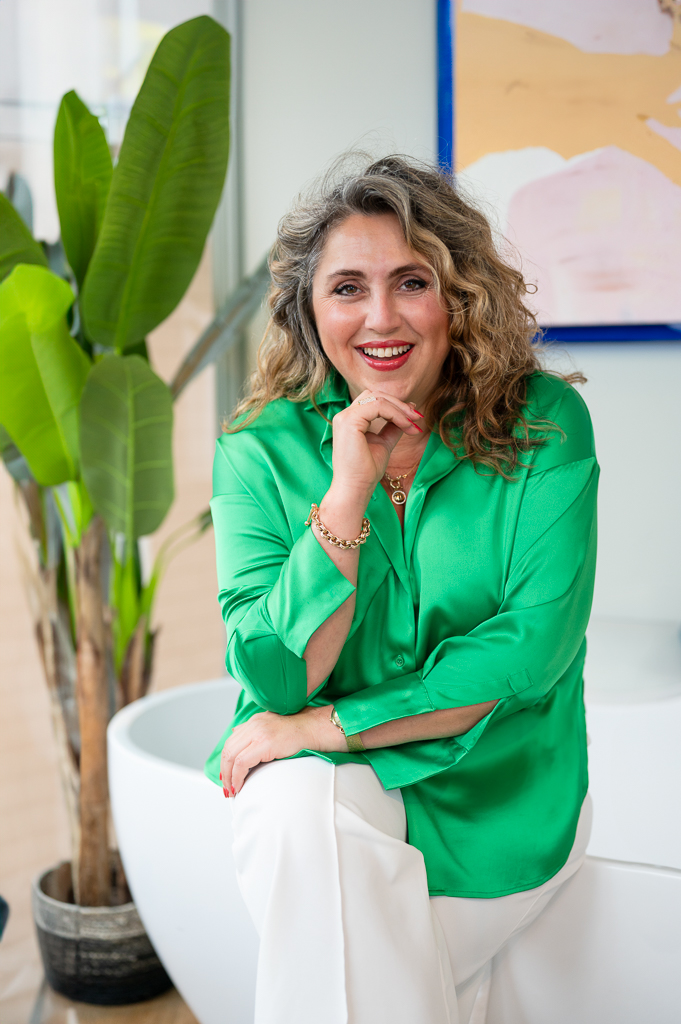 Vrouw met halflang grijs haar met groene blouse en witte pantalon zittend naast een kunst plant